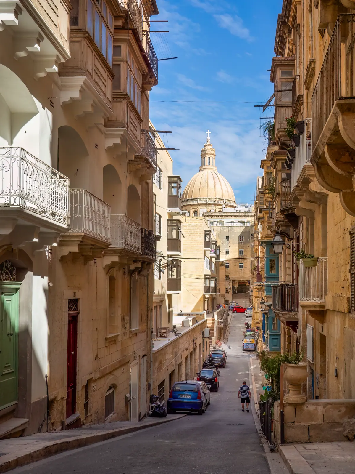 A steep limestone street with balconies and a domed church at the top of the hill.